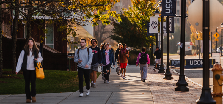 Students walking along a scenic path on the UConn campus with fall foliage in the background.